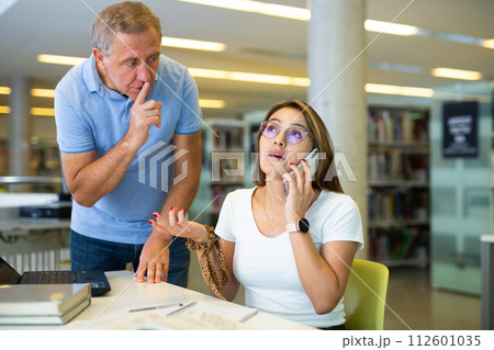 Elderly librarian asks young woman who is talking loudly on mobile phone to keep quiet 112601035