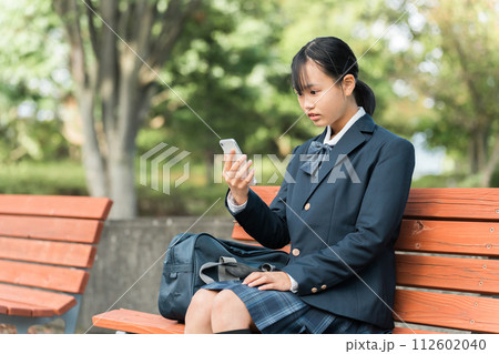 公園のベンチに座りスマートフォンで動画・SNSを見て驚く中学生・高校生の制服姿の女の子 112602040