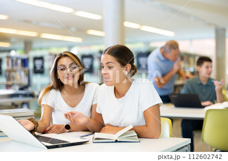 Two friendly adult female students studying in university library, using books and laptop while preparing for lecture Two friendly adult female students studying in university library, using books and laptop while preparing for lecture 112602742