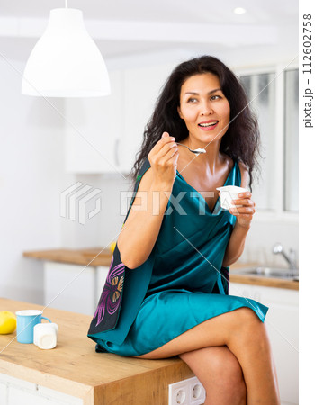 Portrait of positive woman in nightdress eating yogurt at home Portrait of positive woman in nightdress eating yogurt at home 112602758