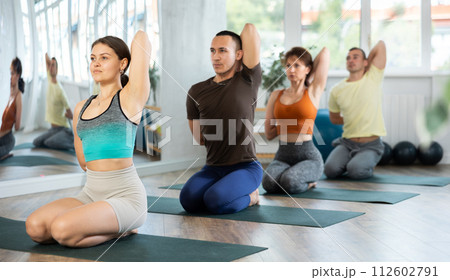Group of sporty people exercising yoga class making Gomukhasana Cow Face Pose in modern fitness center. Focus on female Group of sporty people exercising yoga class making Gomukhasana Cow Face Pose in modern fitness center. Focus on female 112602791