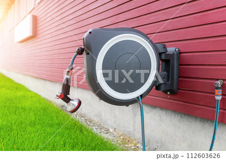 Wall Mounted Retractable Hose Reel. Modern garden lawn watering tip hose with sprinkler hanged at home wall. Lawn and plant tree smart irrigation water system equipment tool Wall Mounted Retractable Hose Reel. Modern garden lawn watering tip hose with sprinkler hanged at home wall. Lawn and plant tree smart irrigation water system equipment tool 112603626