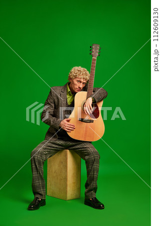 Passionate musician. Curly young man in a suit sitting with guitar against green studio background. Live music event Passionate musician. Curly young man in a suit sitting with guitar against green studio background. Live music event 112609130