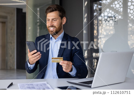 Successful financial transaction and growth. A smiling and satisfied young businessman is sitting at the desk in the office, holding a bank credit card and looking at the phone screen. 112610751