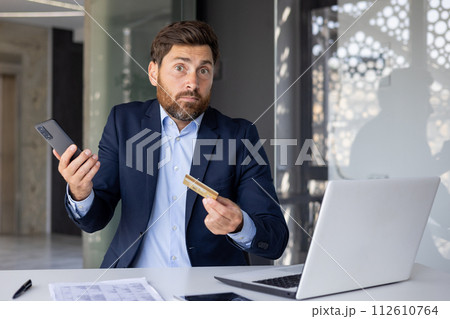 Portrait of a young businessman sitting in the office at the desk, holding a mobile phone and a credit card in his hands, looking disappointed and confused at the camera. Portrait of a young businessman sitting in the office at the desk, holding a mobile phone and a credit card in his hands, looking disappointed and confused at the camera. 112610764