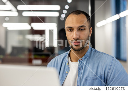Close-up photo of a young hispanic man in a blue denim shirt, sitting at a desk in an office and looking seriously at a laptop screen. 112610978