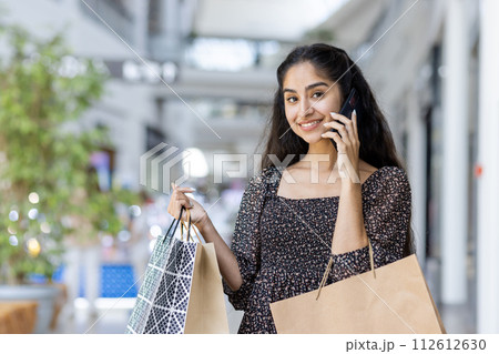Indian young smiling woman standing in a shopping mall, holding paper shopping bags, talking on the phone and looking at the camera. Indian young smiling woman standing in a shopping mall, holding paper shopping bags, talking on the phone and looking at the camera. 112612630