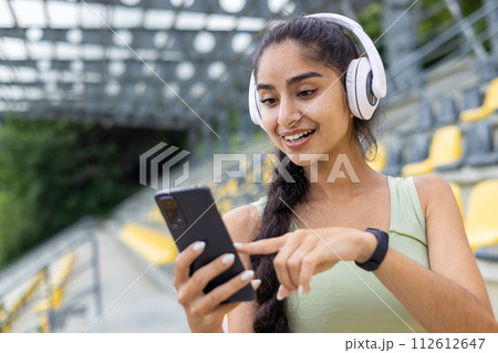 Close-up photo of a young smiling Indian woman in headphones doing sports at a stadium and using a mobile phone. 112612647