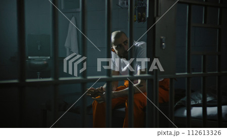 Criminal in orange uniform sits on bed in correctional facility 112613326