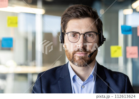 Focused professional wearing a headset and glasses, posing in an office with sticky notes in the background. Represents customer service and corporate environment. 112613624