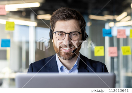 A focused businessman in a suit works on a laptop with a headset in a contemporary office environment. A focused businessman in a suit works on a laptop with a headset in a contemporary office environment. 112613641