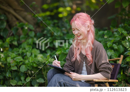 Cute pink hair girl drawing on digital tablet in garden, Woman Doing Freelance Work in Garden, woman with digital tablet with artwork. Cute pink hair girl drawing on digital tablet in garden, Woman Doing Freelance Work in Garden, woman with digital tablet with artwork. 112613823