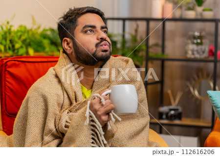 Sick Indian man wrapped in plaid sit alone shivering trembling from cold on chair drinking hot tea Sick Indian man wrapped in plaid sit alone shivering trembling from cold on chair drinking hot tea 112616206