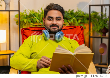 Indian man reading interesting book turning pages smiling enjoying literature taking a rest on chair Indian man reading interesting book turning pages smiling enjoying literature taking a rest on chair 112616208