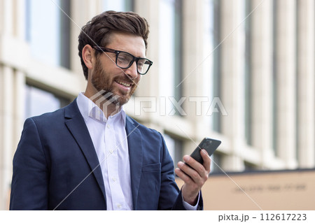 Close-up photo of a smiling young man businessman, lawyer, politician wearing a suit and glasses standing outside an office building, holding a phone and looking at the screen. 112617223