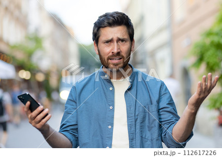 Portrait of an angry and disappointed young man standing on the streets of the city in casual clothes, holding a mobile phone, looking at the camera and spreading his hands. 112617226