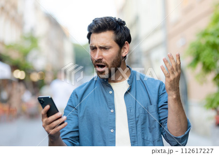 An angry young man in casual clothes stands on a city street and looks worriedly at the phone screen, waving his hands incoherently. 112617227
