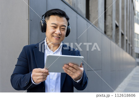 Professional male in a suit multitasking with a digital tablet and headphones outside a modern building. Professional male in a suit multitasking with a digital tablet and headphones outside a modern building. 112618606