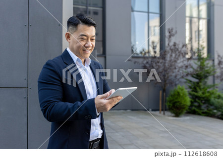 Smiling Asian male professional in a suit engaging with a tablet PC on a sunny day outside a corporate building. Smiling Asian male professional in a suit engaging with a tablet PC on a sunny day outside a corporate building. 112618608