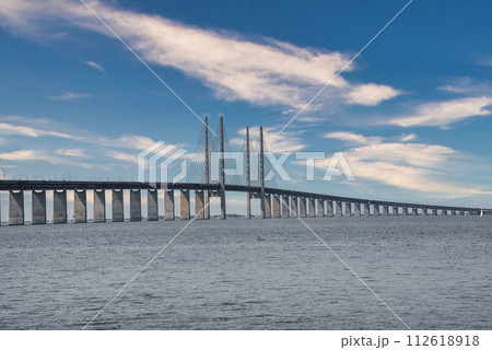 The Oresund Bridge, connecting Copenhagen, Denmark, with Malmo, Sweden, arches over calm waters, its high pylons and cables creating a striking silhouette against a clouddotted sky. The Oresund Bridge, connecting Copenhagen, Denmark, with Malmo, Sweden, arches over calm waters, its high pylons and cables creating a striking silhouette against a clouddotted sky. 112618918