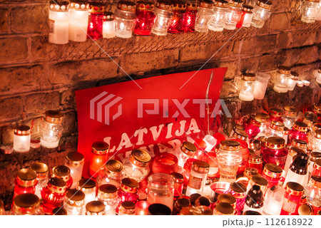 A warm display of candles in red glass jars with golden rims, reflecting a soft glow against a rustic brick backdrop and a red LATVIJA sign, likely celebrating Latvia's Independence Day. 112618922