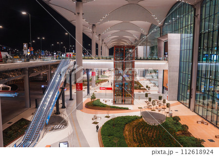 Overlooking Istanbul Airport's interior with white wave arches, soft lights, glass facade, escalator, metro signs, glass lift, seats, and plants. 112618924