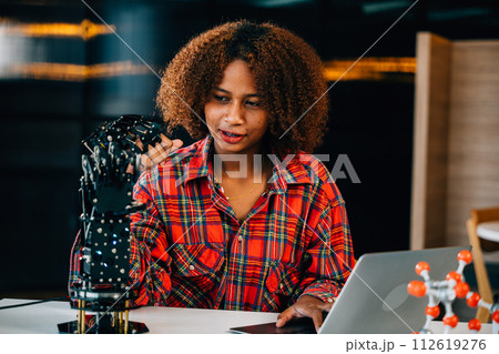 In a classroom a Black teenager studies a robotic arm for her engineering project. Displaying intelligence and skill she controls the arm embracing innovation in technology and science. 112619276