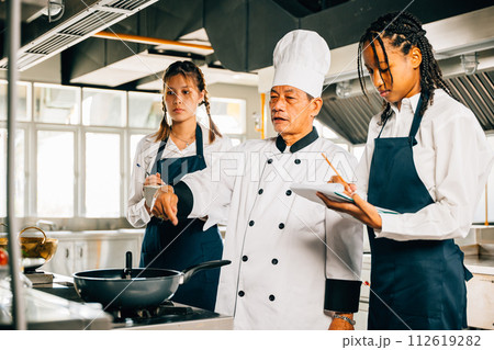 Asian senior chef in uniform educates multiracial students in a restaurant kitchen. Focus on teamwork learning and note-taking in this professional teaching environment. Food Edocation 112619282