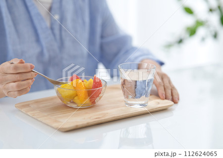 水のグラスを手にフルーツを食べるミドル女性の手元 112624065