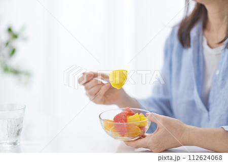 水のグラスを手にフルーツを食べるミドル女性の手元 112624068