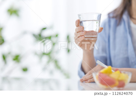 水のグラスを手にフルーツを食べるミドル女性の手元 112624070