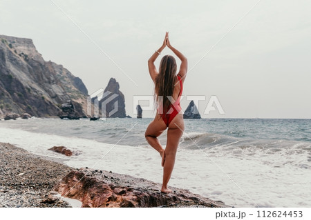Woman sea yoga. Back view of free calm happy satisfied woman with long hair standing on top rock with yoga position against of sky by the sea. Healthy lifestyle outdoors in nature, fitness concept. 112624453