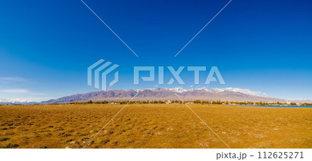 field covered with dry grass bumps with small Kyrgyz town Balykchy and high mountains on the horizon, wide angle panoramic view 112625271