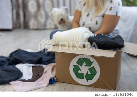 A woman with a dog sorts her clothes while sitting on the floor at home. A woman puts clothes she doesn't wear in a box to be sent for recycling. Clothes recycling concept. 112631040