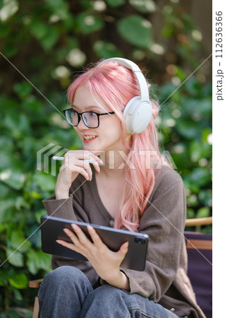 A cute pink-haired girl drawing on a digital tablet and wearing headphones in the garden, a Woman Doing Freelance Work in the Garden, and a woman with a digital tablet with artwork. 112636366