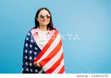 Young smiling happy woman wears casual clothes holding american flag and looking to the camera isolated on blue color background. 112641278