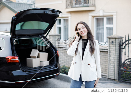 Young business woman standing at the car with parcels, coming home by car. The girl is standing and talking on mobile phone near the car. Concept of buying goods online and delivering them home 112641326