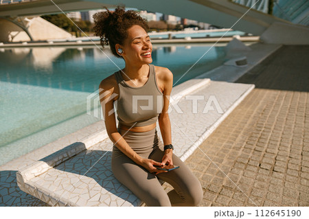 A young woman in sportswear is resting after a workout outside while sitting with smartphone A young woman in sportswear is resting after a workout outside while sitting with smartphone 112645190