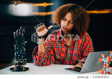 Portrait of a Black teenager studying a robotic arm in a classroom for an engineering project. Skillfully controlling the arm she embraces innovation intelligence and technology. 112645682