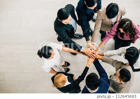 A top view shows four diverse businesspeople forming a circle stacking their hands. This embodies the concepts of unity teamwork and global collaboration in the corporate world. 112645683