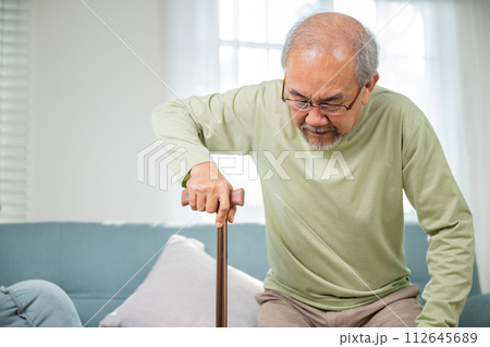Elderly man suffering from knee pain ache holding handle of cane, senior old man typing to stand up from sofa alone with walking cane stick to walk at home, retirement medical healthcare concept 112645689