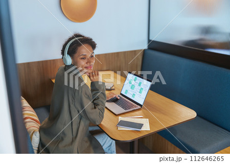 A woman at a table with a laptop, headphones on, using a personal computer A woman at a table with a laptop, headphones on, using a personal computer 112646265