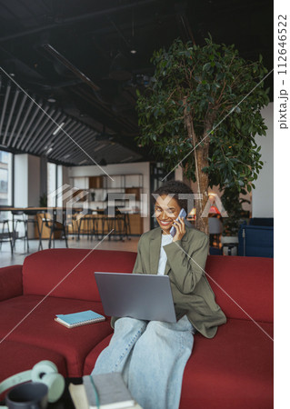 Woman multitasking on a couch with a laptop and cell phone in nice modern coworking office 112646522