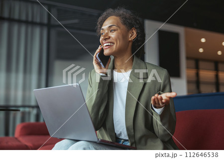 a woman is sitting on a couch talking on a cell phone while using a laptop computer in nice modern coworking office 112646538