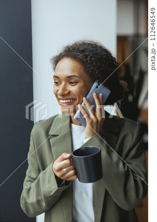 A woman smiles while holding a cup of coffee and talking on a mobile phone 112646549