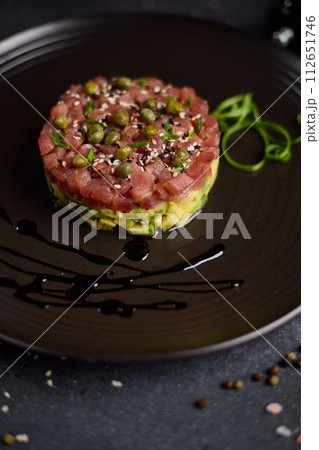 Tuna and avocado tartare with sesame seeds and capers on a dark ceramic plate 112651746