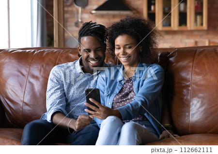 Attractive African couple sit on sofa looking at cellphone screen Attractive African couple sit on sofa looking at cellphone screen 112662111