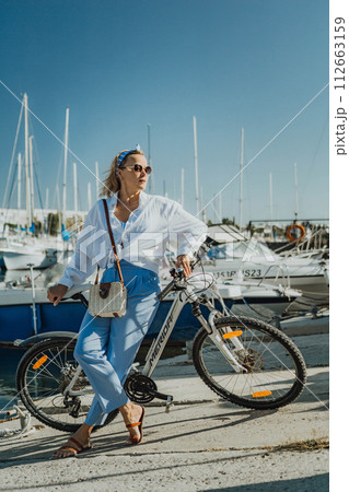 Woman enjoys bike ride along waterfront, Marina surroundings. She is wearing a white shirt and blue jeans, and she has a handbag with her. Capturing outdoor bike ride by waterfront. 112663159