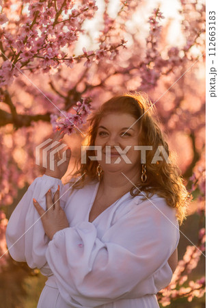 Woman blooming peach orchard. Against the backdrop of a picturesque peach orchard, a woman in a long white dress enjoys a peaceful walk in the park, surrounded by the beauty of nature. 112663183