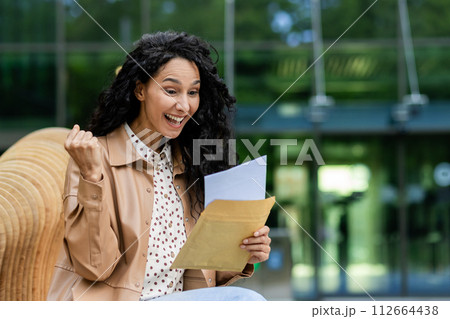 Joyful woman sitting outside, fist pumping while reading excellent news in a letter, expressing excitement and happiness. 112664438